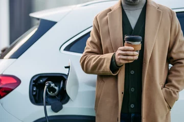 Fotobehang Close up of businessman waiting while electric car is charging, leaning against vehicle, drinking coffee. An electric vehicle charging station. © Halfpoint