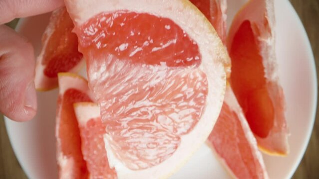 A man's hand takes a wedge of grapefruit and brings it close to the camera. Top view, close-up.