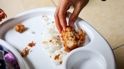 Woman's hand holding Fried chicken with rice placed on a white child's dining table, the food is...