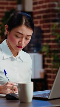 Vertical video Asian worker sulking in office, having frown on face, feeling annoyed by having too much work to do. Employee writing down info on notepad, gloomily solving tasks late at night