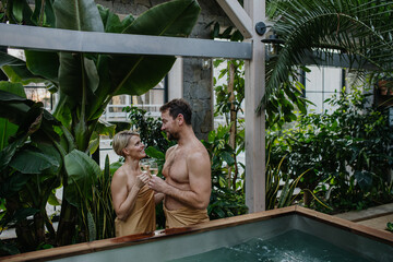 Beautiful couple standing hot tub, toasting, drinking champagne, enjoying romantic wellness weekend...