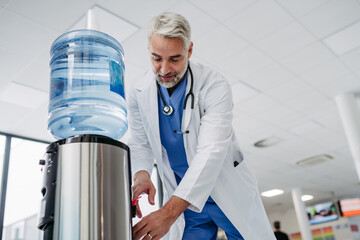 Handsome doctor taking a break during work shift at hospital, drinking water from water dispenser...