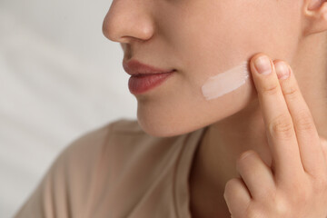 Young woman with dry skin applying cream onto her face on blurred background, closeup