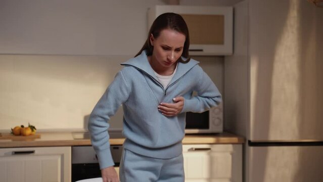 A woman clad in a blue hoodie is clutching her chest in pain, possibly indicating a sudden onset of discomfort or a health concern, while standing in a residential kitchen