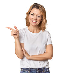 Blonde middle-aged Caucasian woman in studio smiling cheerfully pointing with forefinger away.