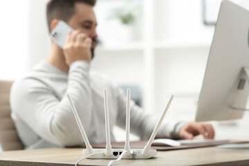 Man talking on smartphone while working at wooden table indoors, focus on Wi-Fi router