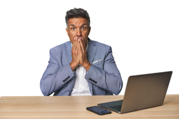 Elegant businessman at desk with laptop shocked, covering mouth with hands, anxious to discover something new.