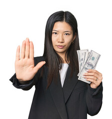 Young Chinese woman with dollars in suit standing with outstretched hand showing stop sign, preventing you.