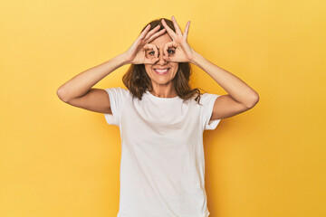 Middle-aged caucasian woman on yellow showing okay sign over eyes