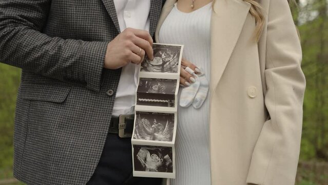 A couple stands close, with the man holding ultrasound photos over the woman's pregnant belly, both radiating parental love and expectation, suitable for stock videos on pregnancy and family.