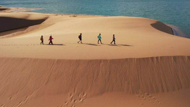 Group of young hikers with backpacks walks at desert dunes and big blue lake. Gobi desert, Mongolia