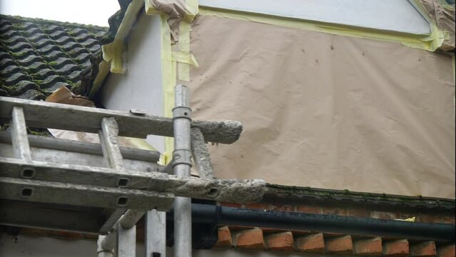 Portable scaffolding with attached ladders being moved away from a prepared work area, namely a roof dormer window, masked off with paper and tape, ready for rendering of the exposed parts.