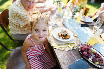 Portrait of a beautiful little girl sitting at table eating grilled food outdoors. Girl at family...
