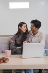 Asian couple watching movies on the internet using laptop at home Smiling Thai man and woman sitting on sofa, hugging each other, looking at computer screen. Surf the web together copy space