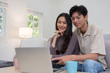 Asian couple watching movies on the internet using laptop at home Smiling Thai man and woman sitting on sofa, hugging each other, looking at computer screen. Surf the web together copy space