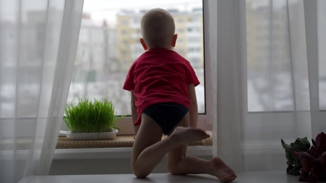 3-year-old child sitting by the window in winter, little boy looks out the window onto the street.
