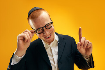 Portrait of smiling young Jewish man in yarmulke, glasses and suit raising finger up against yellow...
