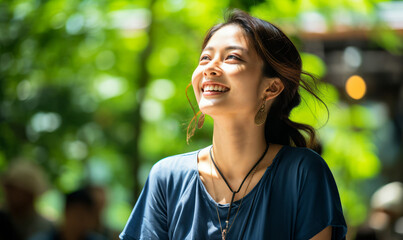 Joyful Asian woman with a bright smile wearing a blue top enjoying a sunny day in a lush green park, radiating happiness and vitality - obrazy, fototapety, plakaty