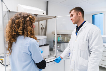 Male doctor and female nurse in lab coats and gloves discuss over a laminar flow hood in a hospital laboratory.