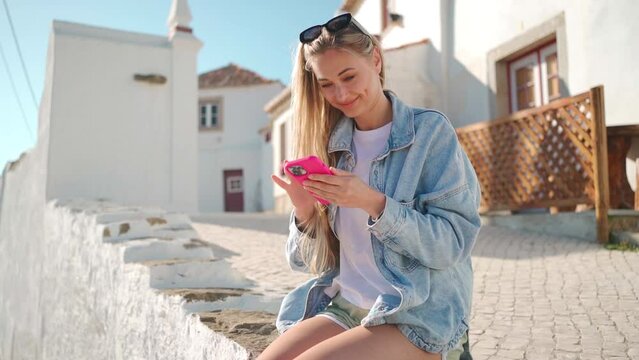 Smiling young woman using smartphone sitting against white houses in Portugal. Beautiful blond female tourist in denim shirt reading message or social media. Happy holiday concept.