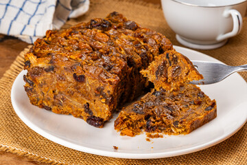 Using metal fork picking a piece of dried fruitcake up from a white ceramic plate on sack cloth on wooden table.
