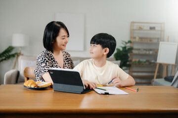 Homeschool Asian young little boy learning online and does homework by using computer and tablet with mother help, teach and encourage. son smile to study at home together with mom