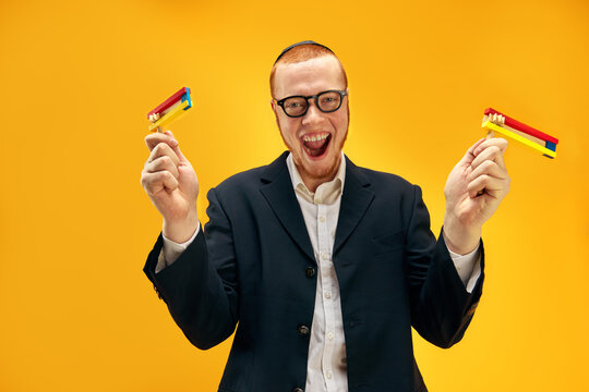 Young redhead Jewish man in glasses, yarmulke playing with wooden noisemaker, gragger against yellow studio background. Concept of Purim holiday, Jewish traditions, history and culture