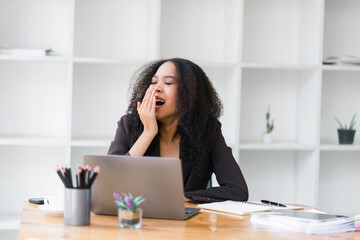 Beautiful Afro American businesswoman using a laptop and yawning while working in a white office.