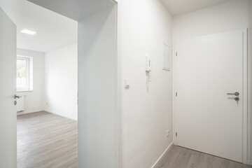An empty room with white walls, a closed entrance door, and an opening leading to a naturally lit room. There is a light switch and intercom by the door.