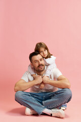 Obraz na płótnie Canvas Cheerful man sitting cross-legged with little girl resting on his back against pink pastel background. Concept of International Day of Happiness, childhood and parenthood, positive emotions. Ad
