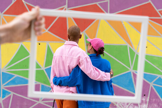 Cropped hand holding picture frame with friends facing towards colorful wall