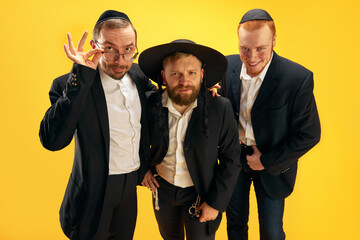 Three Jewish men, friends in traditional Jewish attributes smiling, posing against yellow studio...