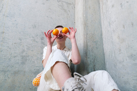 Non-binary person holding oranges over face sitting in front of wall