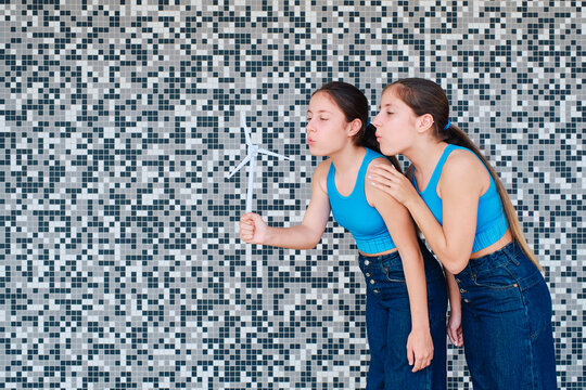 Sisters blowing on wind turbine model in front of textured wall