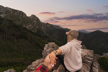 Photography in follow me style. A girl sits on a cliff in the Tatra Mountains and admires the sunset while holding her boyfriend’s hand. - obrazy, fototapety, plakaty