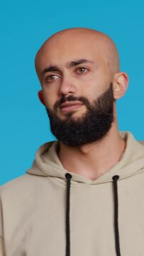 Vertical Video Middle eastern serious adult posing on studio backdrop, feeling confident and smiling over turquoise background. Arabic person acting casual in a beige hoodie, handsome guy. Camera 2.
