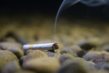 a cigarette with burning embers and ash at the end that emits white smoke between the rocks