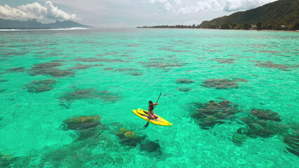 Aerial drone shot woman paddle boarding coral reef tropical island. Young female rows sitting on yellow paddleboard in crystal turquoise sea water. Outdoor lifestyle travel on summer holiday vacation.