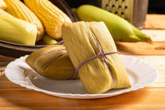 Pamonha and green corn, Pamonha on a white plate and beautiful ears of green corn on rustic wood, selective focus.
