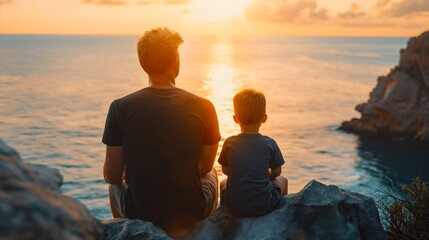 Rearview photography of a father and son sitting together on a rock or cliff on the sea coast, wearing black t shirts and looking at the ocean waves during the golden hour sunset sky, vacation - obrazy, fototapety, plakaty