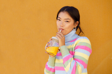 Woman drinking orange juice standing in front of yellow wall