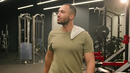 Happy satisfied Latino man athlete walk go to start workout exercise with towel on shoulder finish training fit sport guy fitness trainer instructor male bodybuilder sportsman in gym drinking water