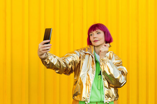 Smiling senior woman taking selfie with smart phone in front of yellow wall