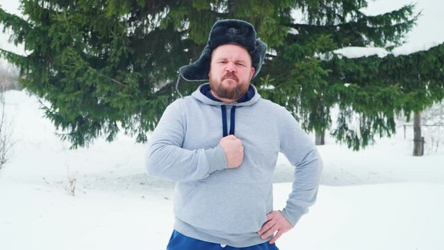 A Russian man with a beard and wearing a winter hat with earflaps beats his chest. Close-up of a Russian man.