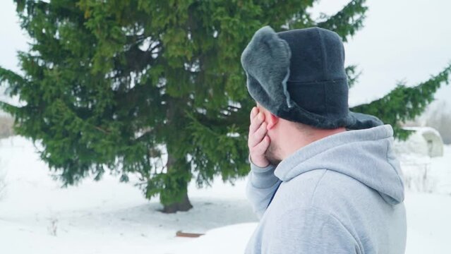 A Russian man scratches his beard, thinking about whether to cut down a tree or not. A man in a winter hat with earflaps. A fir tree grows near the house.