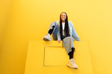 Smiling young woman sitting on concrete block near yellow wall