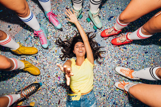 Happy woman gesturing lying on confetti amidst legs of of friends wearing roller skates