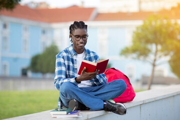 Obraz premium Happy Black Young Man Student In Eyeglasses Reading Book Outdoors