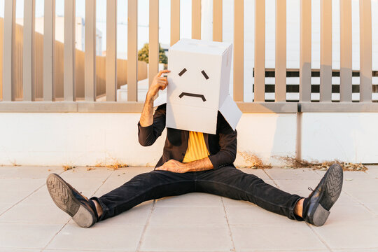 Thoughtful man wearing box with sad face sitting on footpath