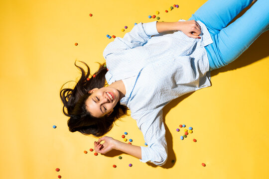Happy woman lying amidst colorful candies against yellow background